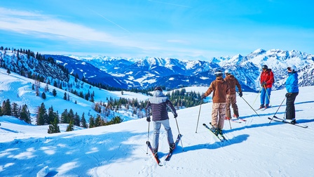 Rückenansicht von fünf Personen in Skianzügen mit Helmen, Ski und Skistöcken, die auf einer Piste stehen. Ihnen gegenüber sind schneebedeckte Berge. Der Himmel ist blau