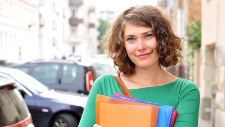 Halbporträt einer Person mit kinnlangen Haaren, die vor ihren Oberkörper mehrere Mappen hält. Links neben der Person sind parkende Autos. Dahinter sind Gebäude