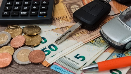 Nahaufnahme mehrerer Euroscheine, Euromünzen, eines Taschenrechners, eines Autoschlüssels, eines Spielzeugautos und eines Stiftes.