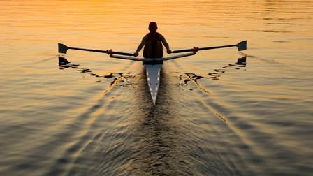 Person mit Kappe im Dämmerlicht des Sonnenuntergangs rudert mit zwei Paddeln in spitz zulaufendem Ruderboot auf Gewässer