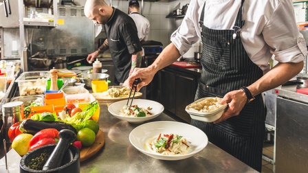 Detailansicht auf Teller in einer Gastronomieküche, ringsum kochende und Essen zubereitende Personen