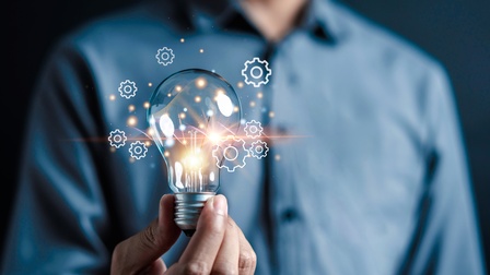 Eine Person in einem blauem Hemd hält eine Glühbirne in die Kamera. Neben der Glühbirne sind Grafiken von Zahnrädern