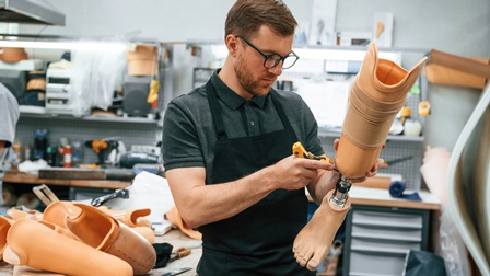 Bärtige Person mit Brillen in schwarzer Arbeitsschürze hält Beinprothese in Händen und blickt darauf, daneben Tisch voller Prothesen, im Hintergrund verschwommen Werkbänke