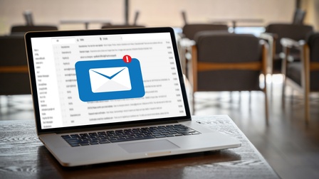 Ein aufgeklappter Laptop steht auf einem Tisch. Auf dem Bildschirm ist ein weißer Briefumschlag auf blauem Hintergrund. Rechts oben ist ein weißes Rufzeichen in einem roten Kreis