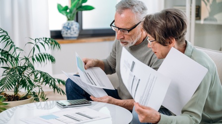 Zwei Personen mittleren Alters mit Brille sitzen auf einer Couch und betrachten Unterlagen, die sie in der Hand halten und die auf einen Beistelltisch liegen, daneben liegt ein Taschenrechner, im Hintergrund zeigt sich ein Wohnraum