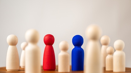 Aufgestellte Spielfiguren aus Holz, von denen 2 rot und blau sowie im Fokus sind. Sie stehen auf einem braunen Holztisch