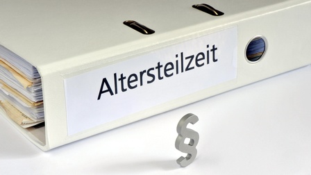 Nahaufnahme eines liegenden, hellen Ordners auf weißem Untergrund. Auf der Seite des Ordners steht in schwarzer Schrift Altersteilzeit. Im Ordner sind Unterlagen. Vor dem Ordner ist ein graues Objekt in Form eines Paragraphenzeichens stehend platziert