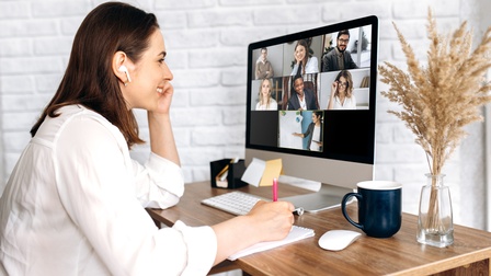 Seitliche Aufnahme einer Person mit Kopfhörern, die an einem Schreibtisch sitzt und auf einen Bildschirm eines Computers blickt, auf dem mehrere Personen online an einer Besprechung teilnehmen. Die Person notiert etwas auf einem Notizblock