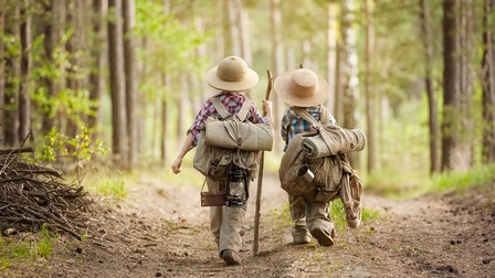 Rückenansicht zweier Kinder mit Rucksäcken, Sonnenhüten und Stöcken die einen Waldweg entlang gehen
