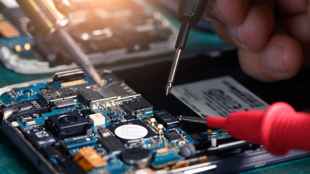 Nahaufnahme einer geöffneten elektronischen Baugruppe. Zwei Messspitzen berühren Bauteile auf einer Leiterplatte. Mehrere elektronische Komponenten, Steckverbindungen und Mikrochips sind auf der Leiterplatte