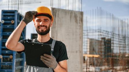Porträt einer Person mit Helm, Handschuhen, Bart und Klemmbrett in der linken Hand. Hinter der Person sind mehrere Betonpflöcke und lange Metallstangen, die aus dem Boden ragen