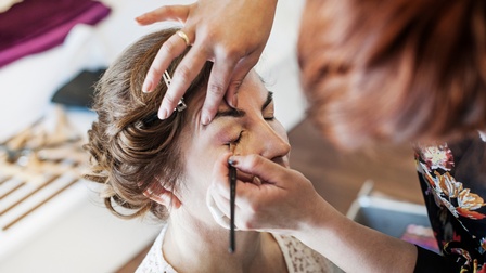 Einer sitzenden Person mit geschlossenen Augen werden von einer anderen Person die Lider geschminkt. In den Haaren hat die sitzende Person eine Haarklammer