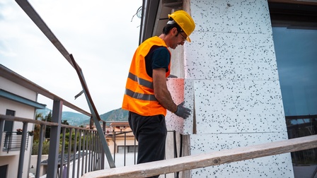 Person in oranger Warnweste, mit gelbem Schutzhelm und Handschuhen bringt Wärmedichtung an Hausfassade an