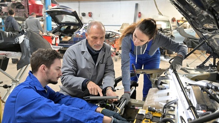 Zwei Lehrlinge arbeiten an Automotor, andere Person in grauem Mantel deutet auf Tablet