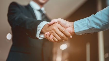 In der linken Bildhälfte ist eine Person in einem Anzug, die einer anderen Person die Hand schüttelt