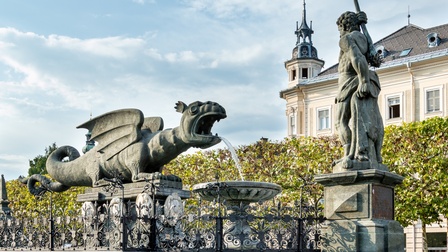 Ansicht eines steinernen Drachens der Wasser speiht, gegenüber stehend steinerner Mann mit Bärenfell um die Hüften und erhobener, hinter Rücken hängender Keule vor historischem Gebäude.