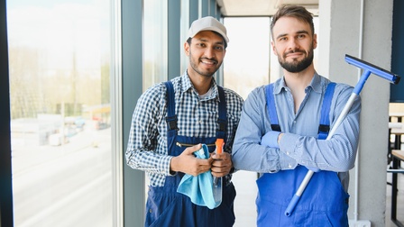 Porträt zweier nebeneinander stehender Personen. Die rechte Person hält einen länglichen Stab mit einem horizontalen Kopf. Die linke Person hält ein blaues Tuch und eine Sprayflasche. Links neben den Personen ist eine Fensterfront