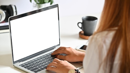 Eine Person tippt auf der Tastatur eines Laptops am Schreibtisch. Daneben stehen eine Dekopflanze, eine Kafeetasse und ein Smartphone.