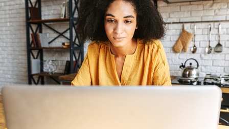 Frontale Aufnahme einer Person mit lockigem, dunklem Haar, die an einem Tisch in einer Küche sitzt und auf einen aufgeklappten Laptop blickt. Im Hintergrund sind ein Herd und weitere Küchenutensilien