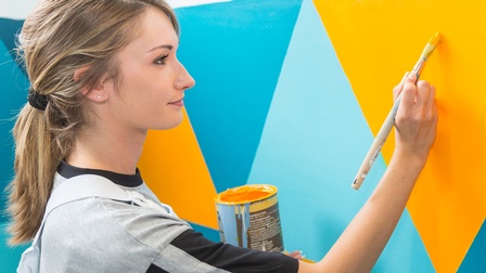 Person steht an Wand und malt gelbblaues Karomuster darauf