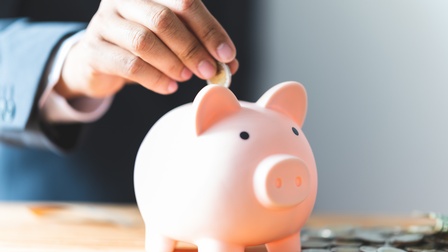 Hand wird Euromünze in ein rosa Sparschwein während weitere Münzen am Tisch verteilt liegen