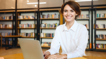 Porträt einer lächelnden Person mit kurzen, braunen Haaren und hellem Hemd, die an einem Tisch sitzt. Vor ihr steht ein aufgeklappter Laptop. Im Hintergrund steht ein langes Bücherregal mit Büchern an der Wand