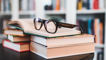 Im Vordergrund liegen auf einem Beistelltisch ein paar Bücher übereinander. Auf dem Stapel liegt ein aufgeklapptes Buch, auf dem eine Brille liegt. Im Hintergrund ist ein Bücherregal mit Büchern