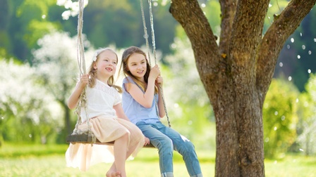 Kinder sitzen auf einer Schaukel, die bei einem Baum befestigt ist, im Hintergrund zeigt sich in der Unschärfe eine grüne und weiße Frühlingslandschaft