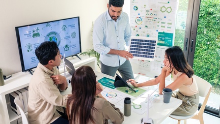 Drei Personen sitzen um einen Tisch. Eine Person steht daneben. Neben ihnen ist ein Bildschirm. Vor ihnen liegen Zettel. Hinter ihnen steht ein großes Plakat. Auf allen drei steht ESG. Am Bildschirm steht außerdem use renewable energy