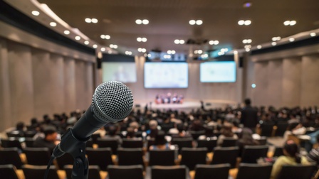 Im Vordergrund ist ein Mikrofon. Im Hintergrund ist ein Saal mit mehreren Sesselreihen, in denen teilweise Leute sitzen. Am Ende des Raums ist eine Bühne, auf der fünf Personen nebeneinander sitzen. Dahinter sind Bildschirme