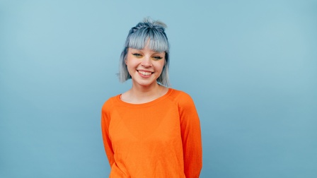 Halbporträt einer Person in orangem Oberteil, gelb-blauen Lidschatten und blauen Haaren vor blauem Hintergrund. Die Arme sind hinter dem Rücken.