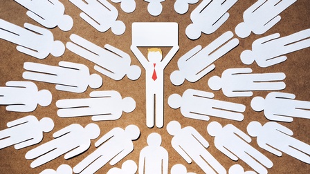 Weiße Papierschnitte von Symbolen von Männern liegen auf einem Tisch. Sie sind in drei Reihen kreisförmig platziert, in der Mitte ist ein Mann-Symbol mit roter Krawatte und blonden Haaren, die herausstechen.  Er hält ein weißes Schild hoch. 