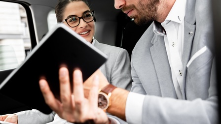 Zwei Personen sitzen in Anzügen in einem Auto nebeneinander. Die rechte Person hält ein Notizbuch. Die linke Person hält Zettel. Die Personen reden miteinander.