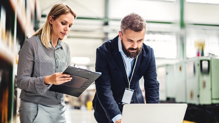 Zwei Personen stehen vor einem niedrigem Tisch in einer Halle. Die linke Person hält ein Clipboard in den Händen und blickt darauf. Die rechte Person beugt sich über den Tisch und hat ihre Hände auf dem vor ihr stehenden, aufgeklappten Laptop