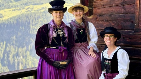 Drei Personen tragen traditionelle Trachten und stehen bzw. sitzen vor einer Holzhütte. Im Hintergrund sind Berge und Wald zu sehen. Alle drei lächeln in die Kamera, die Kleidung ist farblich aufeinander abgestimmt und detailreich verziert.