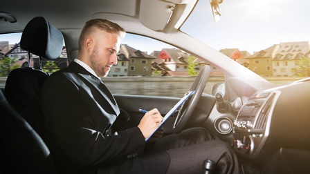 Person sitzt am Fahrersitz eines Autos und notiert auf einem Clipboard