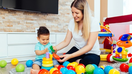 Ein Kleinkind sitzt auf einer grauen Decke und hält in der rechten Hand einen grünen Bauklotz. Direkt daneben sitzt eine erwachsene Person am Boden. Vor ihnen liegen viele bunte Bälle. Hinter ihnen sind weitere Spielsachen