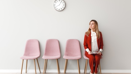 In einem Raum stehen vier Stühle. Auf dem Stuhl ganz rechts sitzt eine Person in einem roten Anzug. Sie hält Unterlagen in den Händen. An der Wand ist eine Wanduhr montiert