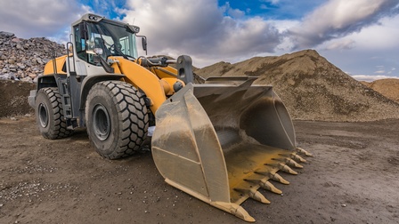 Ein Baufahrzeug mit einer großen Schaufel dran steht auf einem lehmigen Untergrund. Im Hintergrund sind mehrere Hügel aus verschiedenen Rohstoffen wie Steine oder Sand