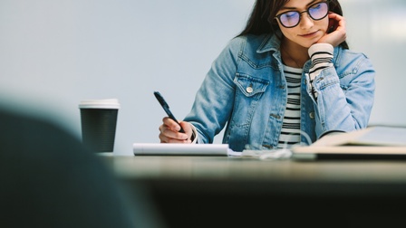Person mit Brillen sitzt an Tisch und blickt darauf, eine Hand im Kinn eingestützt, in anderer Hand Stift auf Notizblock, daneben Becher stehend