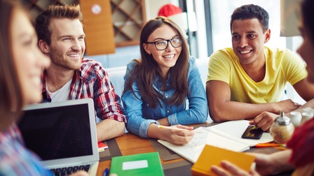 Lächelnde junge Personen an Tisch sitzend mit Notizblöcken, aufgeklapptem Notebook, Stiften, Zuckerstreuer und Smartphone