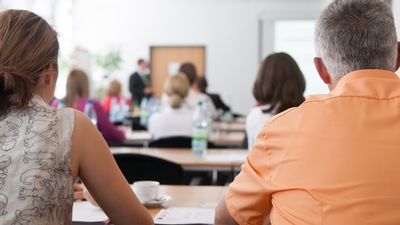 Rückenansicht mehrerer Personen, die in Reihen an Tischen sitzen und nach vorne blicken, wo eine Person steht. Auf den Tischen liegen Unterlagen.