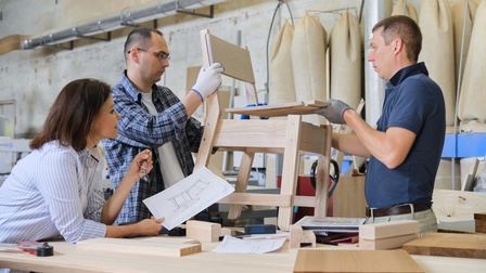 Seitliche Aufnahme dreier neben einem Tisch stehender Personen. Eine Person beugt sich über den Tisch und hält einen Zettel mit der Skizze eines Stuhls. Die anderen beiden Personen halten zwischen sich einen Holzstuhl.