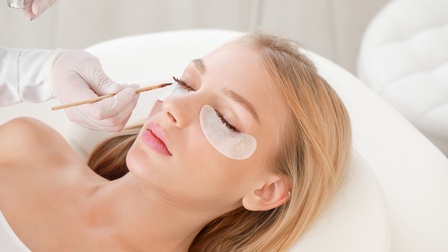 Person mit blonden langen Haaren liegt auf einer Kosmetikliege und ihre Wimpern werden gefärbt