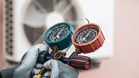 Nahaufnahme der Hände einer Person mit Handschuhen, die ein Objekt halten, an dem an zwei parallelen, schwarzen Leitungen jeweils ein rundes Objekt mit einem Pfeil und einem schwarzen Blatt mit Zahlen drinnen ist