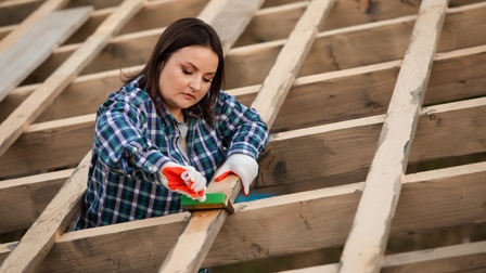 Eine Person mit Handschuhen ist zwischen Holzbrettern eines Regals. Mit der rechten Hand hält sie einen Pinsel an ein Holzbrett, auf den sie auch blickt