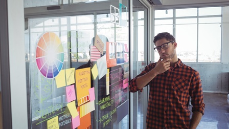Person mit Karohemd und Brille steht nachdenklcih vor einem Whiteboard mit Notizen in einer offenen Glas und Betonräumlichkeit