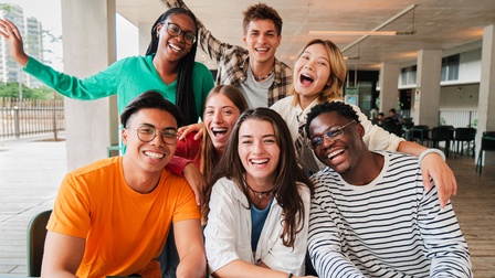 Drei junge Personen sitzen an einem Tisch an einer überdachten Stelle im Freien. Hinter ihnen sind fünf weitere Personen, die sich über die sitzenden Personen lehnen. Jede Person hat den Mund geöffnet und die Mundwinkel nach oben gezogen