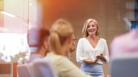 Lächelnde Person mit blonden, schulterlangen Haaren und Businesskleidung in einem hellen Meetingraum stehend, weitere Personen sitzen daneben und blicken in Richtung der vortragenden Person