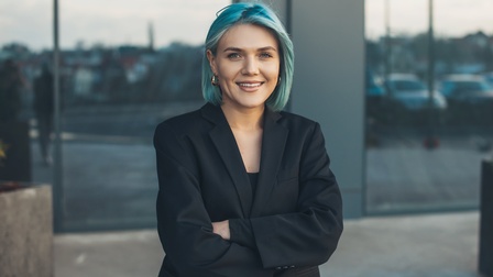 Halbporträt einer Person vor einem Gebäude mit kinnlangen, blauen Haaren und goldenen Ohrringen. Die Arme sind vor der Brust verschränkt und die Person trägt einen schwarzen Blazer.